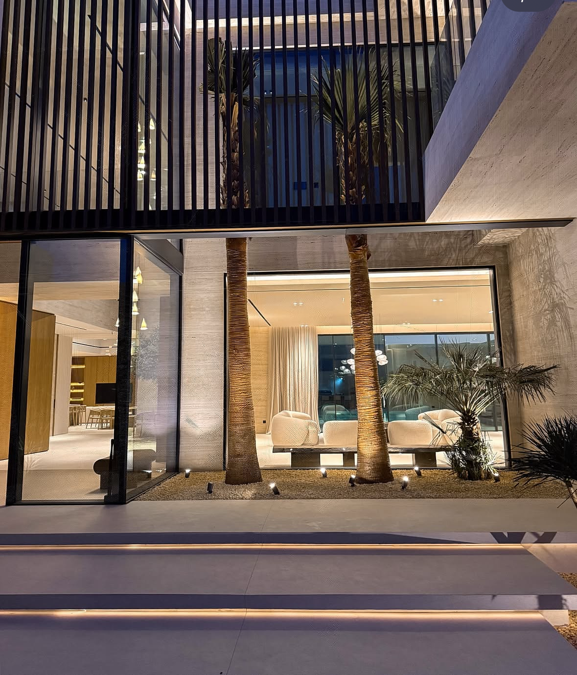 A completed Imperium Group villa at night — travertine walls, timber screening, and palms lit against a glazed double-height living room